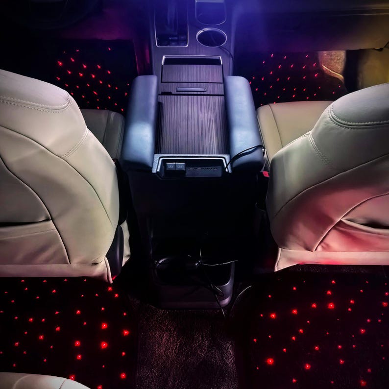 Puede incluir: Interior de coche con asientos de cuero color crema y consola de imitaci&oacute;n madera oscura. Las alfombrillas negras est&aacute;n iluminadas con numerosas luces rojas peque&ntilde;as, creando un efecto de cielo estrellado. Iluminaci&oacute;n ambiental p&uacute;rpura y roja.