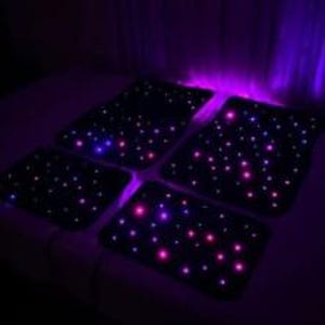 Puede incluir: Cuatro alfombrillas negras para coche iluminadas con peque&ntilde;as luces LED de colores, que simulan un cielo estrellado. Las luces son principalmente rosas, azules y moradas. Las alfombrillas son rectangulares y est&aacute;n dispuestas sobre una superficie oscura.