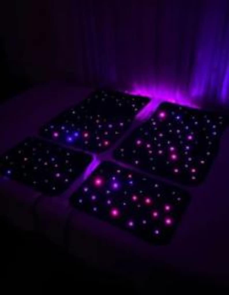 Puede incluir: Cuatro alfombrillas negras para coche iluminadas con peque&ntilde;as luces LED de colores, que simulan un cielo estrellado. Las luces son principalmente rosas, azules y moradas. Las alfombrillas son rectangulares y est&aacute;n dispuestas sobre una superficie oscura.
