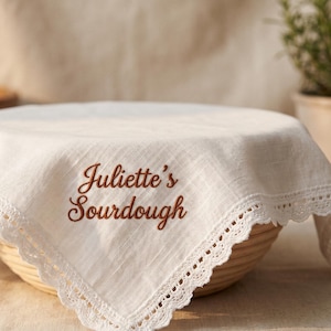 May include: A white linen sourdough bread cover with a scalloped lace edge, embroidered with "Juliette's Sourdough" in brown script. The cover is draped over a round, woven basket. Other wooden bowls and a potted plant are in the background.
