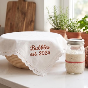 May include: A white fabric bowl cover with a delicate lace trim, featuring the embroidered text "Bubbles est. 2024" in a rust-colored thread. The cover is draped over a round, light-colored woven proofing basket used for bread dough.