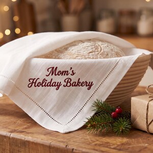 May include: A round, ribbed proofing basket containing a loaf of bread, covered by a white linen cloth with a decorative hem. The cloth features the embroidered text 'Mom's Holiday Bakery' in dark red script, accented by a small pine sprig.