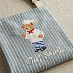 May include: Blue and white striped apron with an embroidered chef bear holding a whisk and bowl. The bear wears a chef's hat, white coat, and blue striped pants. The name "Chef Smith" is embroidered below the bear.
