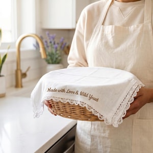 Puede incluir: Una cesta redonda de mimbre cubierta con un pa&ntilde;o blanco con un delicado ribete de encaje. El pa&ntilde;o tiene bordado el texto "Made with Love & Wild Yeast". La cesta se muestra en un entorno de cocina.