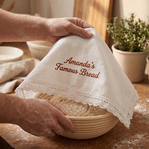 Puede incluir: Una tela blanca para cubrir el pan, con borde de encaje, bordada con "Amanda's Famous Bread" en escritura marr&oacute;n, cubre un pan en una cesta redonda de mimbre. La escena sugiere panader&iacute;a casera.