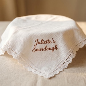 Puede incluir: Una cubierta de pan de lino blanco con un borde de encaje festoneado. Las palabras "Juliette's Sourdough" est&aacute;n bordadas en escritura marr&oacute;n en la esquina. La cubierta est&aacute; drapeada sobre un objeto redondo.