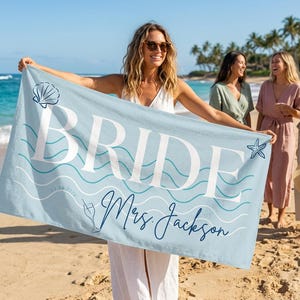 May include: A light blue beach towel with the word "BRIDE" in large white letters. The towel has a seashell and starfish graphic, with the name "Mrs. Jackson" in cursive. The towel is held by a person on a sandy beach.