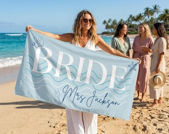 Puede incluir: Una toalla de playa azul claro con la palabra "BRIDE" en letras blancas grandes. La toalla tiene un gráfico de concha y estrella de mar, con el nombre "Mrs. Jackson" en cursiva. La toalla es sostenida por una persona en una playa de arena.