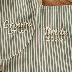 May include: Two striped aprons, one reading "Groom EST 2026" and the other "Bride EST 2026". The aprons are sage green and white striped with beige straps. The text is embroidered in a cursive font.