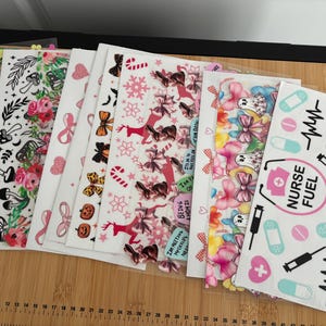 May include: A collection of decorative stickers with various designs, including floral patterns, bows, and holiday-themed graphics. One sticker sheet features medical-themed illustrations with the text "NURSE FUEL". The stickers are arranged on a wooden surface.