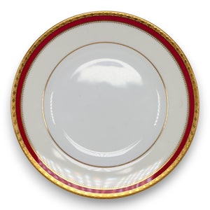 May include: A white ceramic plate with a red and gold border. The plate has a gold rim and a red band with gold detailing. The centre of the plate is white.