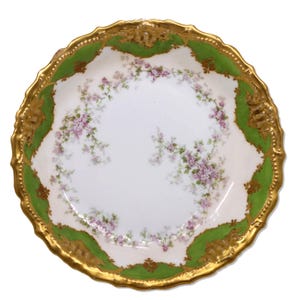 May include: A white porcelain decorative plate with a gold rim and green and gold accents. The plate features a floral design with pink flowers and green leaves. The plate has a scalloped edge.