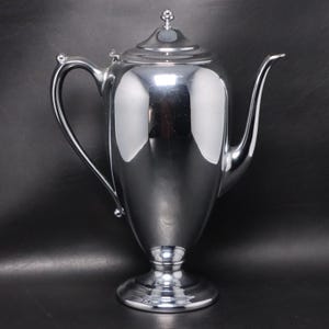 May include: A shiny, silver-colored coffee pot with a curved handle and a long, slender spout. The pot has a lid with a decorative finial and sits on a pedestal base. The polished surface reflects the surrounding environment.