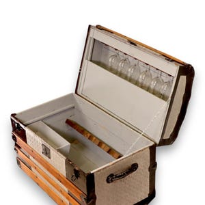 May include: A vintage-style trunk with a light-colored patterned exterior, dark metal corner reinforcements, and brown wooden slats. The interior is painted white, featuring a built-in rack for hanging wine glasses and a divided storage compartment.