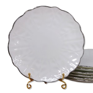 Puede incluir: Plato de cerámica blanco con borde festoneado y borde plateado. El plato tiene una superficie texturizada y se exhibe sobre un soporte de metal dorado. Una pila de platos similares es visible en el fondo.