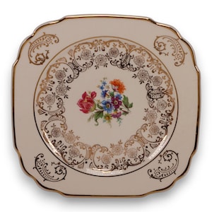 May include: A square, off-white plate with a scalloped edge and gold trim. The plate features a central floral arrangement in red, blue, and orange, surrounded by a gold filigree design. The plate is a vintage piece.