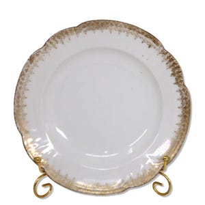 Puede incluir: Plato de porcelana blanca con borde festoneado y ribete dorado. El plato se exhibe sobre un soporte de metal dorado. El ribete dorado presenta un patrón repetitivo alrededor del borde.
