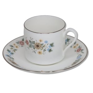 May include: White teacup and saucer set with a floral design. The cup and saucer have a silver trim. The floral pattern includes yellow, pink, and blue flowers with green leaves. The cup is sitting on the saucer.