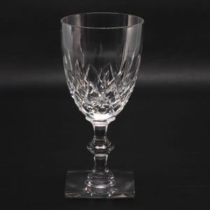 May include: A clear crystal wine glass with a diamond-cut pattern on the bowl. The stem has a decorative design and sits on a square base. The glass is empty and reflects light, set against a dark background.