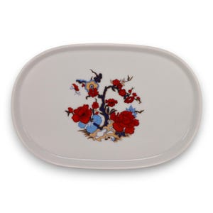 Puede incluir: Plato ovalado blanco para servir con un diseño floral. El diseño presenta flores rojas, detalles azules y un patrón de ramas negras. El plato tiene una forma clásica para servir aperitivos o postres.