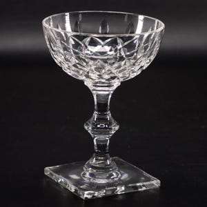 May include: A clear, cut-glass champagne coupe with a square base and a decorative stem. The bowl of the glass features a diamond pattern. The glass is designed for serving champagne or other sparkling wines.