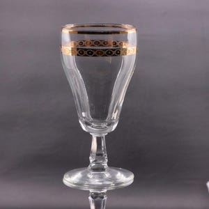 May include: A clear glass goblet with a gold band around the rim and a decorative gold pattern below. The goblet has a flared bowl, a slender stem, and a round base. The glass is transparent, allowing light to pass through.
