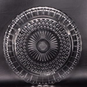 May include: Clear glass serving plate with a detailed pattern. The plate features a central sunburst design, surrounded by rows of small, raised circles. The outer edge has a scalloped rim with a repeating rectangular pattern, creating a decorative and functional piece.