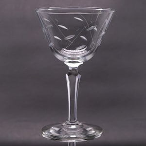 Puede incluir: Una copa de cóctel transparente con base y un cuenco cónico y ancho. La copa presenta un diseño grabado de pequeñas formas de hojas y líneas curvas. El tallo es delgado y la base es un disco plano y circular. Para cócteles.