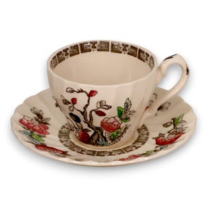 May include: Cream-colored teacup and saucer set with a floral and tree design. The cup has a geometric rim and a curved handle. The saucer has a scalloped edge and a matching pattern. The set is likely made of porcelain.