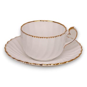 May include: White teacup and saucer set with gold trim. The teacup has a curved handle and a scalloped rim. The saucer has a matching scalloped edge. The set is made of porcelain and is suitable for tea or coffee.