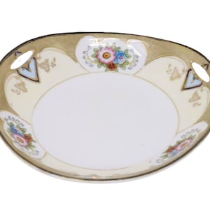 Puede incluir: Un plato ovalado de porcelana con interior blanco y borde crema. El borde está decorado con detalles dorados, motivos florales y formas geométricas azules y doradas. El plato tiene dos asas pequeñas.