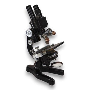 May include: A black metal binocular microscope featuring two eyepieces, a rotating nosepiece with multiple objective lenses, a mechanical stage with adjustment knobs, a coarse and fine focus dial, and a circular mirror at the base.