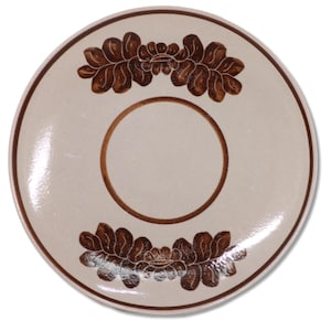 May include: A round, off-white ceramic plate with a brown floral design. The plate has a brown rim and a brown circle in the center. Two brown floral patterns are on the top and bottom of the plate.