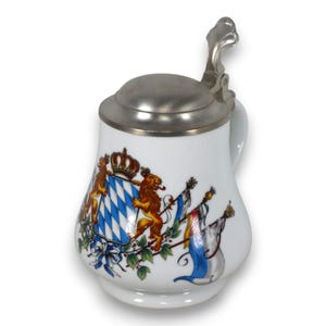Op de afbeelding: Witte keramische bierpul met een scharnierend metalen deksel en handvat. De pul heeft een kleurrijk Beiers wapenontwerp met leeuwen, een kroon en vlaggen. Het ontwerp is in tinten blauw, goud en wit.