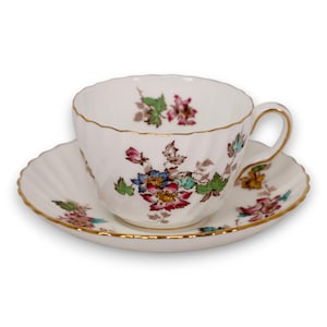 May include: White teacup and saucer set with a floral design in red, blue, and green. The teacup and saucer have a gold trim. The teacup has a handle and a ridged design. The saucer has a scalloped edge.