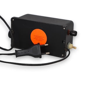 Puede incluir: Un dispositivo eléctrico negro con un cable de alimentación y un enchufe. El dispositivo tiene un dial, una pequeña perilla beige y una pegatina naranja con texto. El dispositivo probablemente se utiliza para aplicaciones eléctricas.