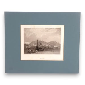 May include: A sepia-toned engraving of a coastal town with mountains in the background, featuring small sailboats and a rowboat with figures in the foreground. The print is mounted on a solid slate blue mat board with the word AJACCIO printed below.