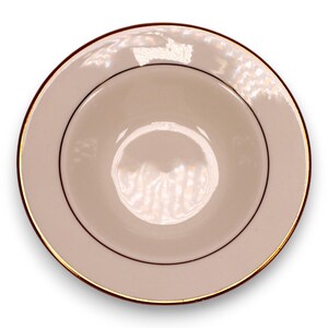 Könnte beinhalten: Eine runde, cremefarbene Keramikschale mit einem goldenen Rand und einer dünnen, dunkelbraunen Akzentlinie. Das Innere der Schale ist in einem helleren Cremeton gehalten, wodurch ein subtiler Kontrast entsteht. Das Design ist schlicht und elegant, geeignet für Suppen oder andere Gerichte.