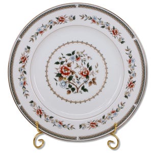 May include: White porcelain plate with a floral design. The plate features a central floral arrangement with red, blue, and white flowers, surrounded by a decorative border. A gold-colored rim and a gold-colored plate stand complete the design.
