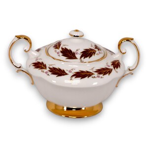 May include: A white porcelain sugar bowl with a lid, featuring a gold base and handles. The bowl is adorned with a pattern of maroon leaves and small purple berries, outlined with gold trim. The lid has a small gold knob.