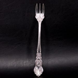 May include: A silver-toned metal cocktail fork featuring an ornate, embossed handle with intricate floral and fruit motifs. The utensil has three slender tines and a decorative base that transitions into a smooth, tapered neck leading to the head.