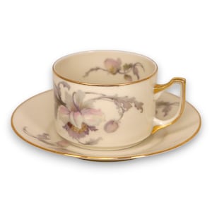 May include: Cream-colored teacup and saucer set with a floral design in shades of purple and gray. The cup and saucer have gold trim. The teacup handle is also trimmed in gold.