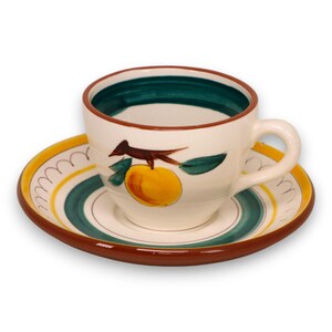 Puede incluir: Un juego de taza y platillo de cerámica. La taza y el platillo son blancos con un diseño pintado a mano de una fruta amarilla, un pájaro marrón y hojas verdes. El platillo tiene rayas amarillas, verdes y marrones.