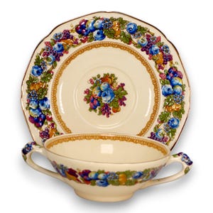 May include: Cream-colored vintage teacup and saucer set. The set features a floral design with blue roses, purple grapes, and other fruits. The cup has two handles and a matching design around the rim. The saucer has a scalloped edge and a gold trim.