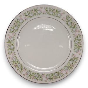 Puede incluir: Plato de cerámica blanco con borde floral. El borde presenta un patrón de pequeñas flores azules, rosas y verdes con hojas verdes. El plato tiene un borde plateado.