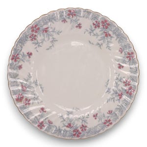 May include: A round white ceramic plate with a scalloped edge and a gold-toned rim. The border features a repeating pattern of small pink flowers with grey-blue leafy vines trailing along the circumference of the plate's wide rim.