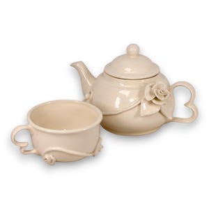 May include: A cream-colored ceramic teapot with a matching teacup. The teapot features a lid, a curved spout, and a sculpted rose detail on the side. The teacup has a curved handle and a small rose accent. Both pieces have a smooth, glossy finish.