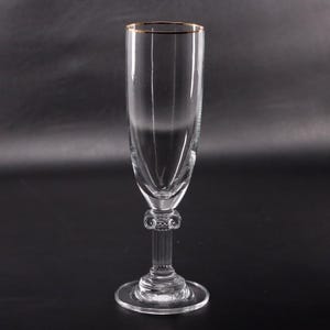 May include: A clear champagne flute with a gold rim. The stem features a decorative design, and the base is round. The glass is tall and slender, perfect for serving sparkling wine or champagne at a special event.