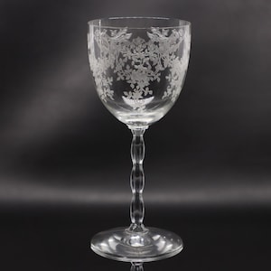 May include: A clear glass goblet featuring an intricate etched floral and leaf pattern around the upper bowl. The stem has a distinct wavy, multi-lobed design leading down to a circular base. The glass is transparent with white etchings.