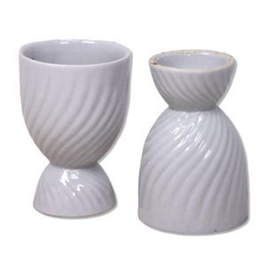 May include: Two white ceramic egg cups with a spiral design. One cup is upright, the other is inverted. The cups have a pedestal base and a wide opening. The design is a classic and elegant addition to any kitchen.